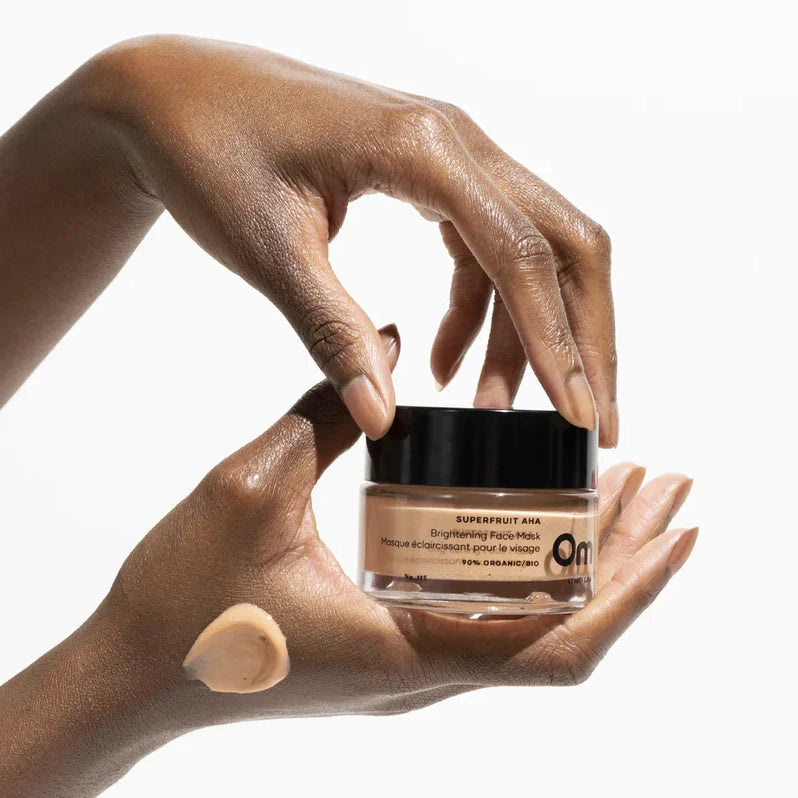 A woman's hand holding a jar of superfruit AHA face mask by Om Organics, with a layer of the mask on the person's hand showing the texture of the mask.