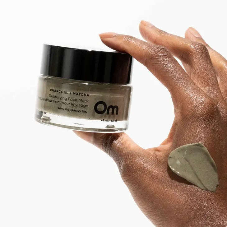 A woman's hand holding a jar of charcoal and matcha detoxifying face mask by Om Organics, with a layer of the mask on the person's hand showing the texture of the mask.