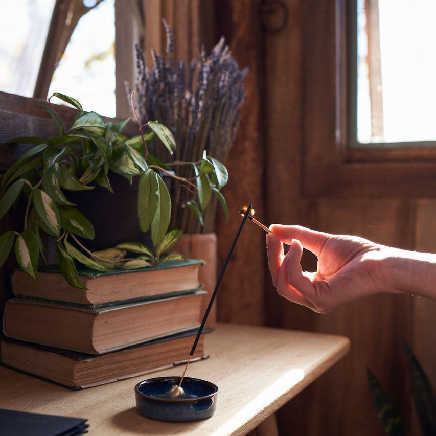 A woman lighting an incense stick by Province Apothecary, in an incense holder, on a desk with lavender, plants, and books in the background.