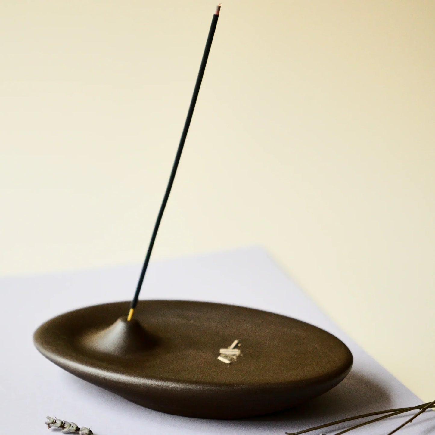 An incense holder in dark brown color, handmade with ceramic stoneware by Siesta Studio, Turkey, holding an incense stick with the ashes having fallen onto the holder.