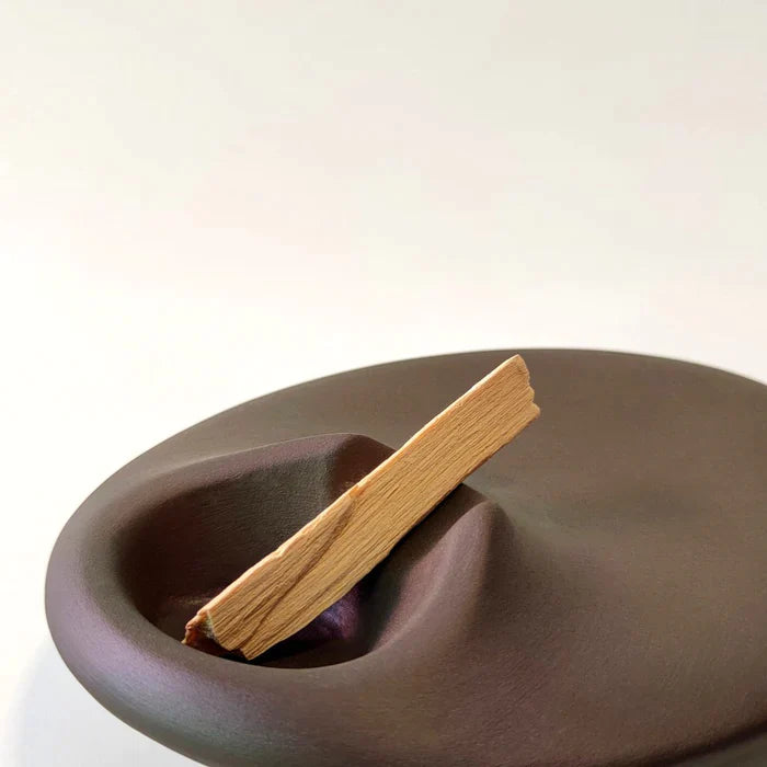 A palo santo holder in dark brown color, handmade with ceramic stoneware by Siesta Studio, Turkey, holding a palo santo stick.